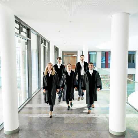 Personen in Richterroben Zwei Frauen und ein Mann in Richterroben laufen in einem Gang entlang auf die Kamera zu, links und rechts des Ganges stehen zwei große weiße Säulen.